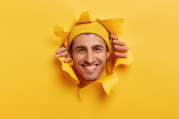 Photo gratuite joyeux homme de race blanche avec un sourire à pleines dents, montre des dents blanches, regarde à travers le trou déchiré dans le papier