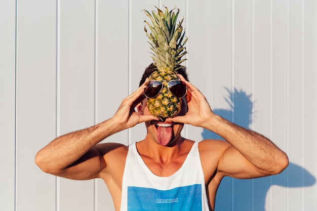 Joyeux homme souriant montrant une langue, tenant un ananas avec des lunettes de soleil
