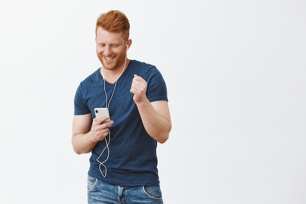Joyeux insouciant beau mâle mature avec des cheveux et des muscles rouges, tenant un smartphone, regardant l'écran avec un large sourire, écoutant de la musique dans des écouteurs