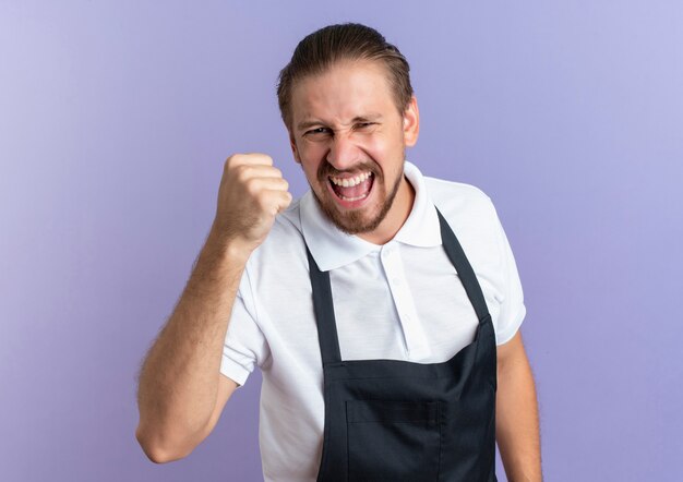 Joyeux jeune beau coiffeur portant l'uniforme levant le poing isolé sur violet