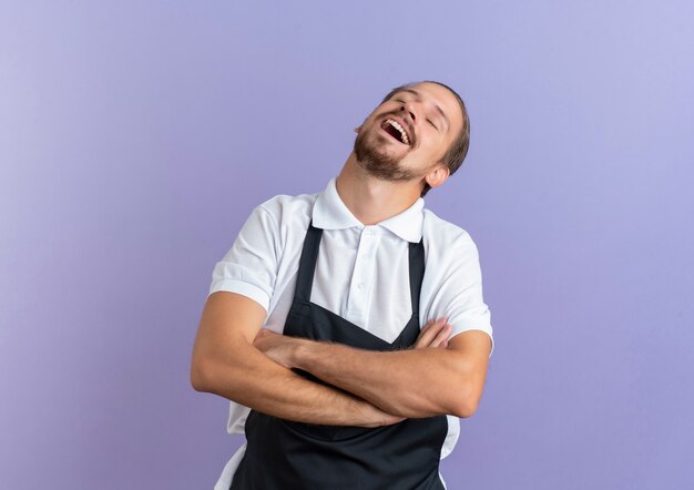 Joyeux jeune beau coiffeur en uniforme debout avec une posture fermée et les yeux fermés isolés sur violet