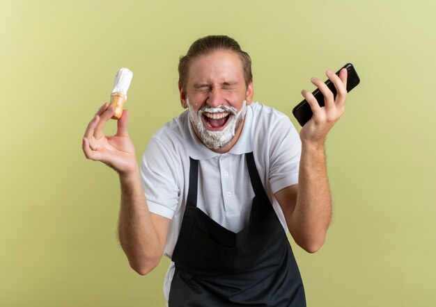 Joyeux jeune beau coiffeur en uniforme tenant un téléphone mobile et un blaireau avec les yeux fermés et avec de la crème à raser appliquée sur sa barbe isolée sur vert olive