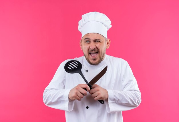 Joyeux jeune beau cuisinier en uniforme de chef faisant un clin d'œil et tenant une cuillère à fentes et un couteau isolé sur un mur rose