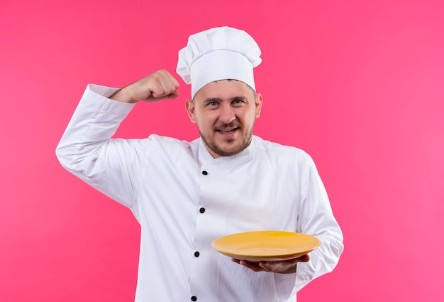 Joyeux jeune beau cuisinier en uniforme de chef tenant une assiette gesticulant fort isolé sur un mur rose