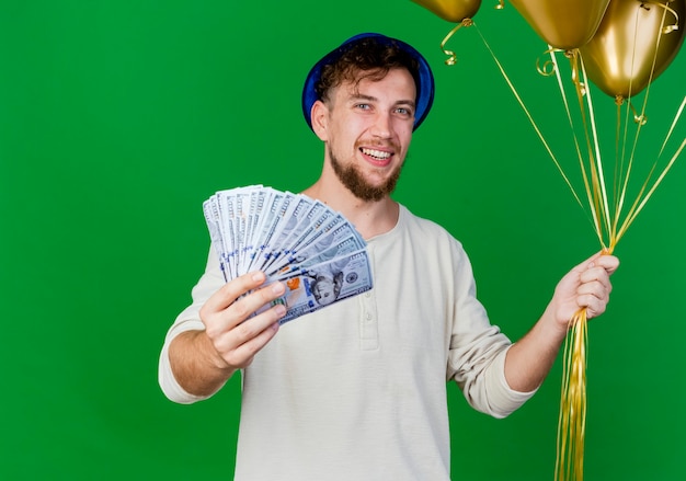 Joyeux jeune beau mec de fête slave portant chapeau de fête tenant des ballons et étirant de l'argent et regardant la caméra isolée sur fond vert avec espace de copie