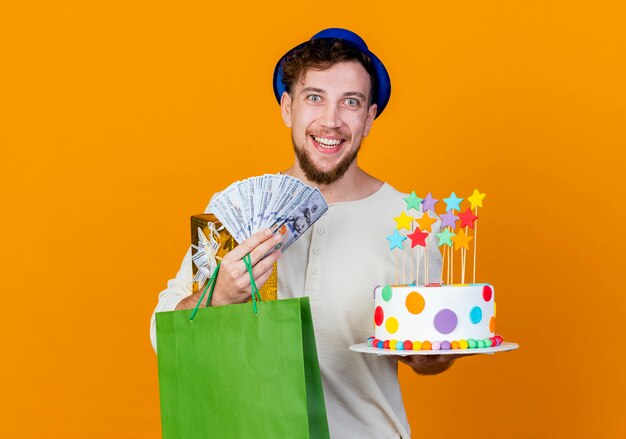 Joyeux jeune beau mec de fête slave portant un chapeau de fête tenant un sac en papier et un gâteau d'anniversaire avec des étoiles regardant la caméra isolée sur fond orange avec espace de copie