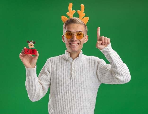 Joyeux jeune beau mec portant bandeau de bois de renne avec des lunettes tenant jouet arbre de Noël avec date regardant la caméra pointant vers le haut isolé sur fond vert
