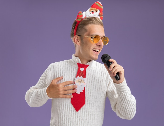 Joyeux jeune beau mec portant bandeau de père Noël et cravate avec des lunettes tenant le microphone en gardant la main sur la poitrine en regardant le chant latéral isolé sur fond violet