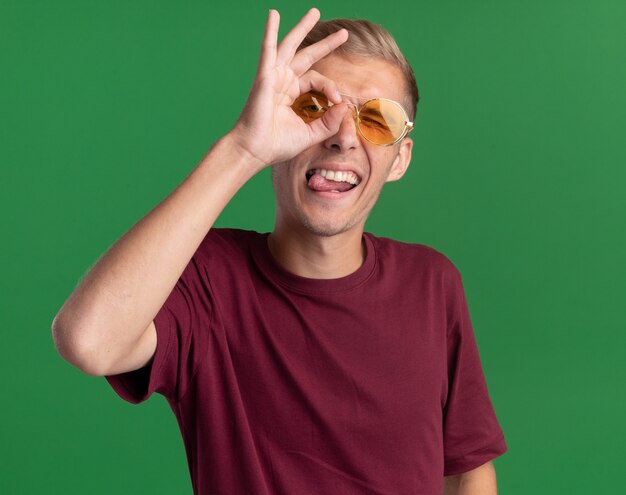 Joyeux jeune beau mec portant une chemise rouge et des lunettes montrant le geste de regard isolé sur le mur vert