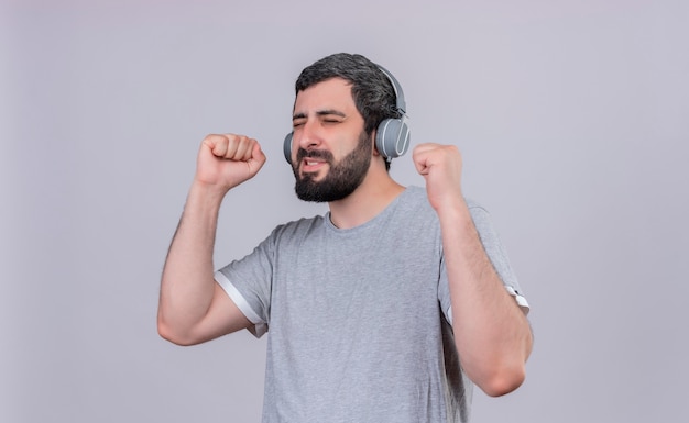 Joyeux Jeune Bel Homme Caucasien Portant Des écouteurs, écouter De La Musique Avec Les Yeux Fermés Et Les Poings Levés Isolé Sur Blanc