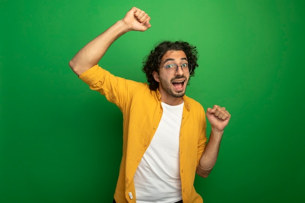 Joyeux jeune bel homme portant des lunettes serrant et levant le poing vers le haut à l'avant isolé sur mur vert