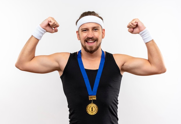 Joyeux jeune bel homme sportif portant un bandeau et des bracelets et une médaille autour du cou gesticulant fort isolé sur mur blanc