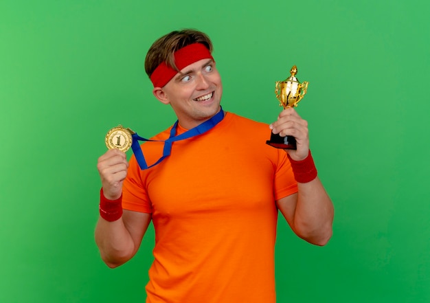 Joyeux jeune bel homme sportif portant bandeau et bracelets avec médaille autour du cou tenant la médaille et la coupe du gagnant en regardant la tasse isolée sur le vert