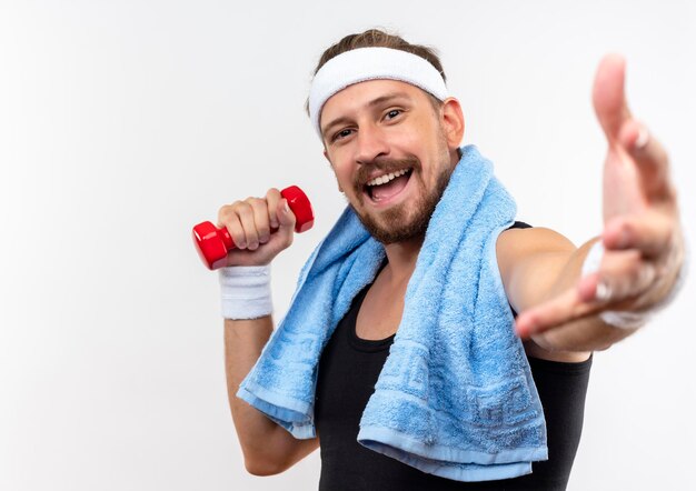 Joyeux jeune bel homme sportif portant un bandeau et des bracelets tenant un haltère et tendant la main vers avec une serviette autour du cou isolé sur un mur blanc