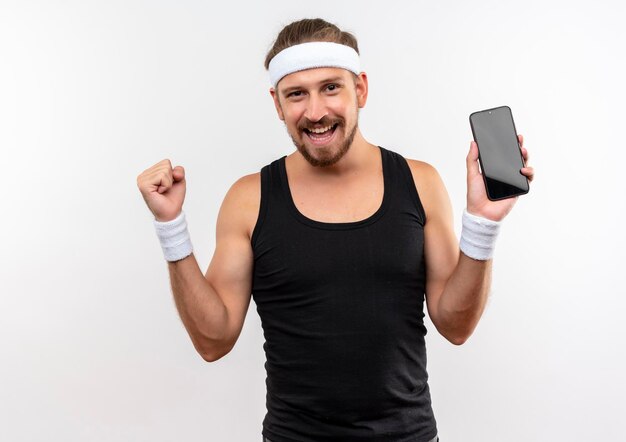 Joyeux jeune bel homme sportif portant un bandeau et des bracelets tenant un téléphone portable et serrant le poing isolé sur un mur blanc