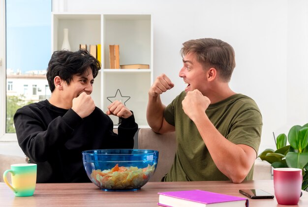 Joyeux jeune blond et brune beaux mecs assis à table en gardant le poing et en se regardant