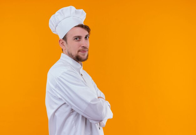 Un joyeux jeune chef barbu homme en uniforme blanc tenant les mains jointes tout en regardant sur un mur orange