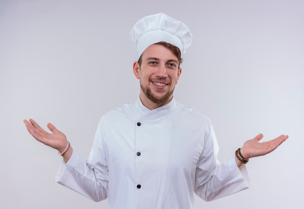 Un joyeux jeune chef barbu homme vêtu d'un uniforme de cuisinière blanc et chapeau ouvrant ses mains tout en regardant sur un mur blanc