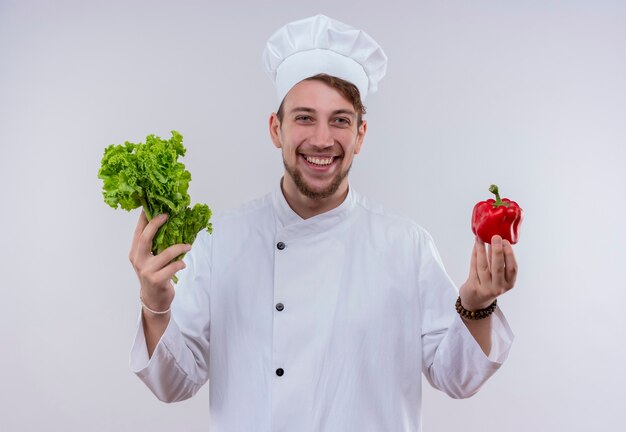 Un joyeux jeune chef barbu homme vêtu d'un uniforme de cuisinière blanc et chapeau tenant la laitue verte et le poivron rouge tout en regardant sur un mur blanc