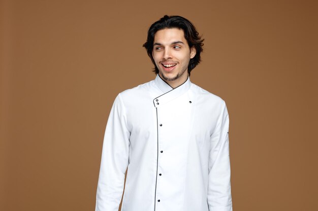 joyeux jeune chef masculin portant l'uniforme regardant le côté souriant isolé sur fond marron