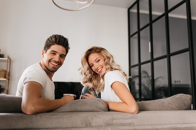 Joyeux jeune couple assis sur un canapé et regardant en arrière. Deux personnes souriantes dans des appartements modernes, buvant du café le matin.