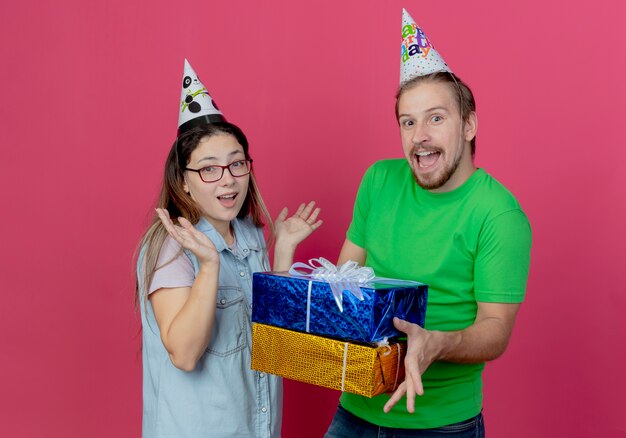 Joyeux jeune couple portant chapeau de fête regarde l'homme détient les coffrets cadeaux et fille lève les mains isolés sur le mur rose
