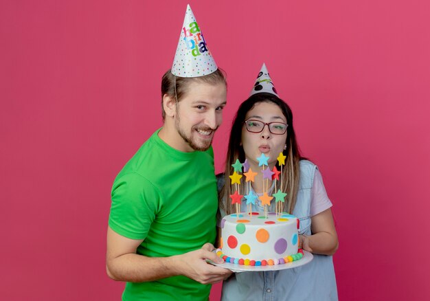 Joyeux jeune couple portant chapeau de fête tient le gâteau d'anniversaire à isolé sur mur rose