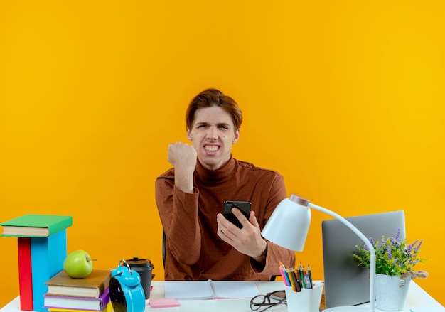 joyeux jeune étudiant garçon assis au bureau avec des outils scolaires tenant le téléphone et montrant oui geste