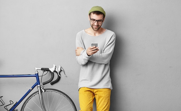 Joyeux jeune hipster caucasien avec chaume habillé avec élégance jouant au jeu vidéo sur le gadget électronique activé sur le Web. Heureux cycliste mec souriant dans des verres surfer sur Internet à l'aide de wifi sur téléphone portable