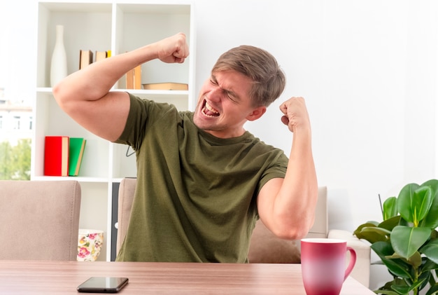 Joyeux jeune homme beau blond est assis à table avec tasse et téléphone levant les poings à l'intérieur du salon