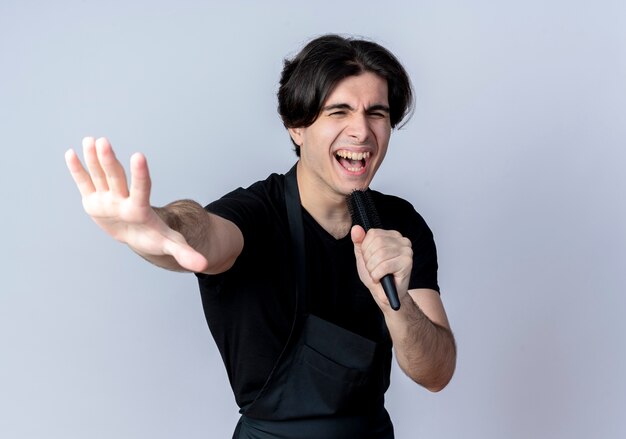 Joyeux jeune homme beau coiffeur en uniforme tenant le peigne et le chant isolé sur fond blanc