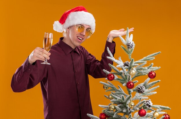 Joyeux jeune homme blond portant bonnet de Noel et lunettes debout près de sapin de Noël décoré pointant vers lui tenant un verre de champagne regardant la caméra isolée sur fond orange