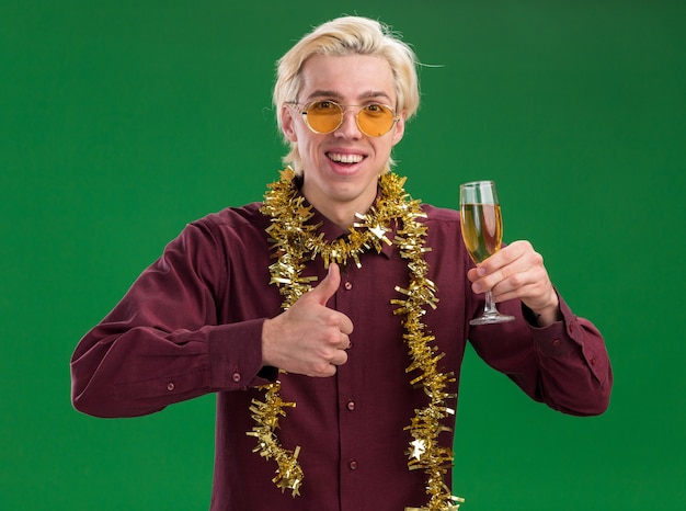 Joyeux jeune homme blond portant des lunettes avec guirlande de guirlandes autour du cou tenant un verre de champagne montrant le pouce vers le haut isolé sur mur vert