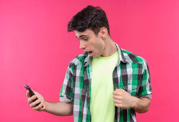 Joyeux jeune homme caucasien portant une chemise verte regardant le téléphone dans sa main et montrant oui geste sur mur rose isolé