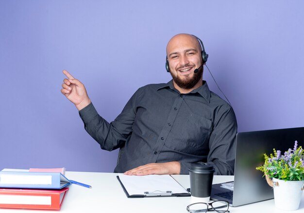 Joyeux jeune homme de centre d'appels chauve portant un casque assis au bureau avec des outils de travail pointant sur le côté isolé sur violet