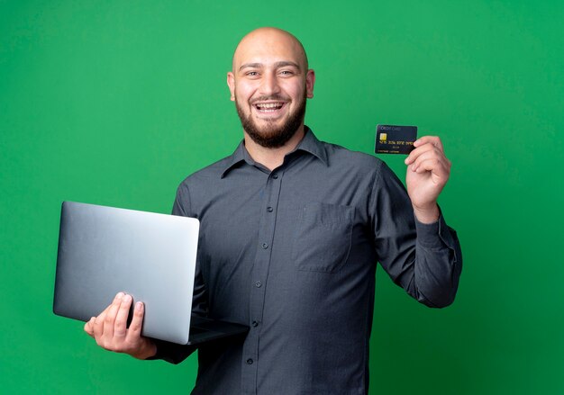 Joyeux jeune homme de centre d'appels chauve tenant un ordinateur portable et une carte de crédit isolé sur vert