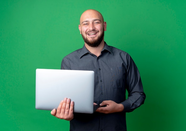 Joyeux jeune homme de centre d'appels chauve tenant un ordinateur portable isolé sur vert