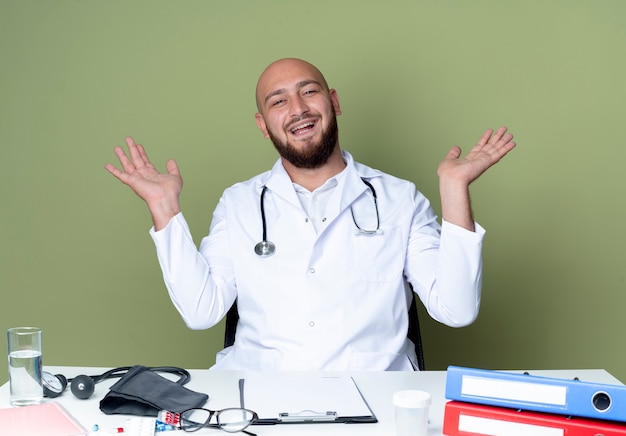 Joyeux jeune homme chauve portant une robe médicale et un stéthoscope assis au bureau