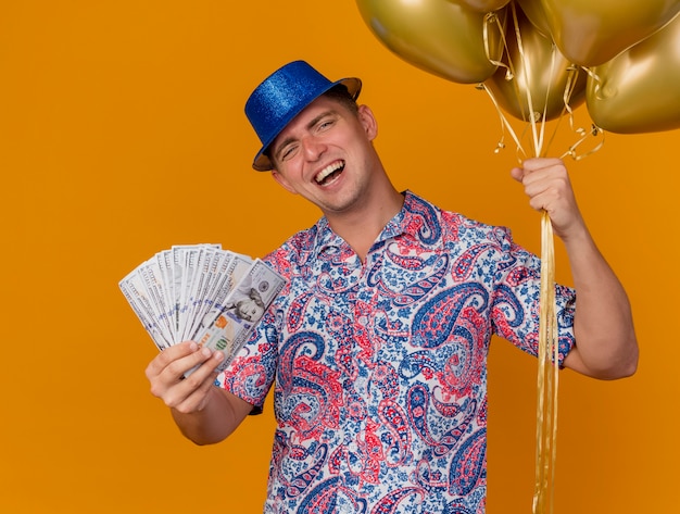 Joyeux jeune homme de fête portant un chapeau bleu tenant des ballons avec de l'argent isolé sur orange
