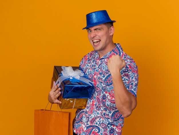 Joyeux jeune homme de fête portant un chapeau bleu tenant des coffrets cadeaux avec sac montrant oui geste isolé sur orange