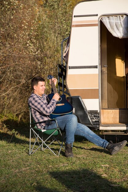 Joyeux jeune homme jouant de sa guitare devant son camping-car rétro dans les montagnes. Homme relaxant dans les montagnes.