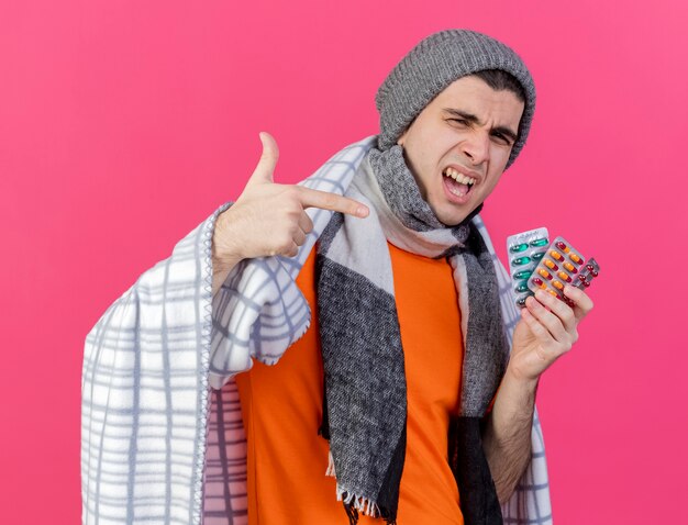 Joyeux jeune homme malade portant un chapeau d'hiver avec écharpe enveloppé dans une tenue à carreaux et des points à des pilules isolés sur rose