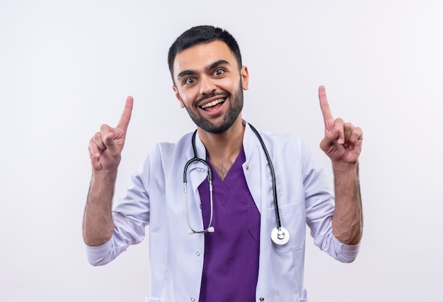 Joyeux jeune homme médecin portant une robe médicale stéthoscope pointe vers le haut sur un mur blanc isolé