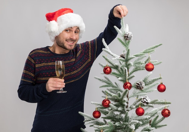 Joyeux jeune homme de race blanche portant un chapeau de Noël debout près de l'arbre de Noël tenant un verre de champagne regardant la caméra saisissant la branche d'arbre isolé sur fond blanc