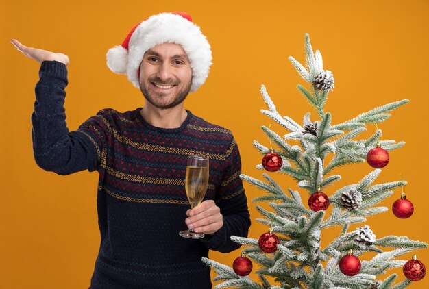 Joyeux jeune homme de race blanche portant un chapeau de Noël debout près de sapin de Noël décoré tenant un verre de champagne montrant une main vide isolée sur un mur orange