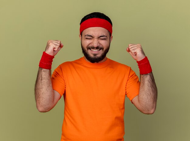 Joyeux jeune homme sportif aux yeux fermés portant bandeau et bracelet montrant oui geste isolé sur fond vert olive