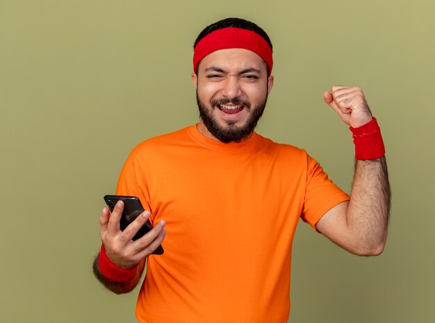 Joyeux jeune homme sportif portant bandeau et bracelet tenant le téléphone et montrant oui geste isolé sur fond vert olive