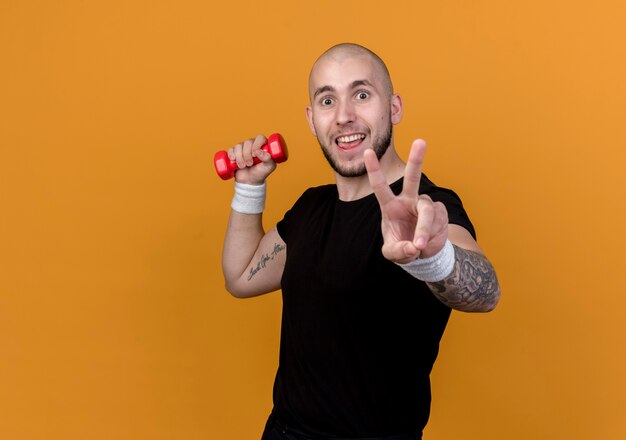 Joyeux jeune homme sportif portant un bracelet tenant un haltère et montrant le geste de paix isolé sur fond orange avec espace de copie