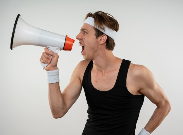 Joyeux jeune homme sportif regardant côté portant un bandeau et un bracelet parle sur haut-parleur isolé sur mur blanc