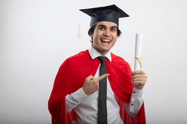 Joyeux jeune homme de super-héros portant cravate et chapeau diplômé tenant et points au diplôme